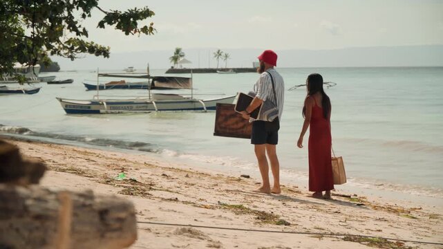 Couple walking sandy beach with tea box and picnic basket, barefoot stroll toward anchored boats, warm golden light, calm turquoise water, gentle breeze, intimate travel ritual, soft footprints