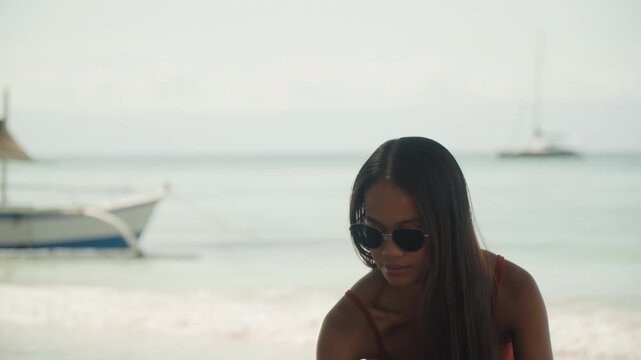 Black woman wearing sunglasses on beach, closeup portrait with braided hair and downward gaze, boats bobbing in calm water behind, sunlit skin and red dress, reflective influencer vibe with fashion