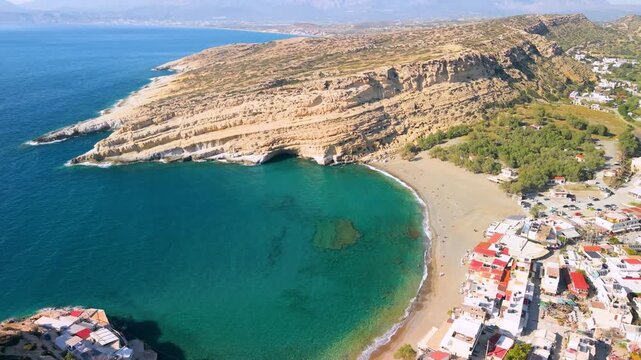 Aerial view of Matala beach with yellow sandstone cliffs, white village and turquoise sea in Crete, Greece. Scenic drone panorama of iconic coastline, sandy shore and Mediterranean houses, 4k.
