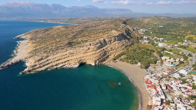 Aerial panorama of Matala coastline, South of Crete. Wide drone view of white village between rugged cliffs, iconic caves and sandy beach. Cinematic landscape of best Cretan beaches in Greece, 4k.