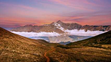 Naklejka premium Mountain landscape above Kazbegi in Georgia with winding trail, low clouds, and dramatic sunset sky.