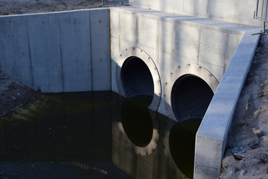 Two Large Round Concrete Drainage Pipe Outlets in Concrete Wall Reflecting in Stormwater Pond