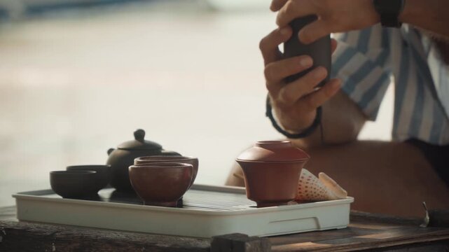 Hands preparing tea set on beach, small clay teapots and cups on wooden tray, slow pouring ritual, soft sunlight, blurred ocean background, calm mindful atmosphere, striped shirt and watch visible