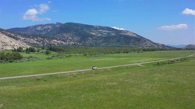 Drone wide shot of small car driving through vast green open landscape with mountains in Kazakhstan, minimalist composition emphasizing scale, isolation, freedom, escape, long distance travel concept