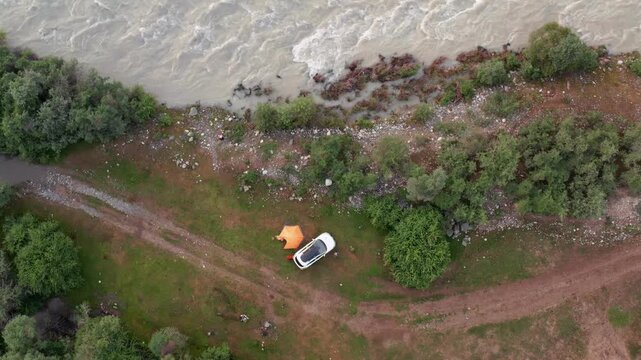 Drone descends and approaches riverside campsite with parked car and tent near Chilik River, revealing top view of shoreline, vegetation and flowing water in Kazakhstan landscape