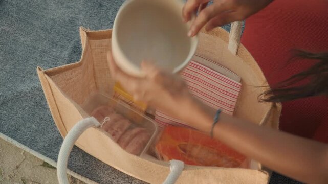 Straw tote packed with picnic essentials, woman reaches into bag to retrieve sealed container and snacks, closeup of hands organizing compartments, red dress sleeve visible, casual beach preparation,