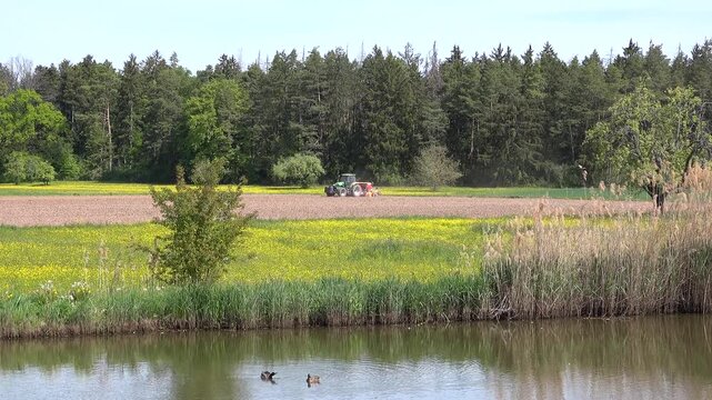 Ein Traktor pfl&uuml;gt einen Acker im Fr&uuml;hling 2026 mit zwei schwimmenden Enten auf einem Teich im Vordergrund