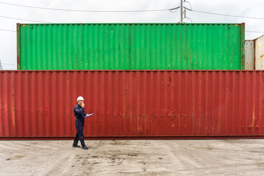 Logistics worker talking on walkie talkie while walking past stacked shipping containers