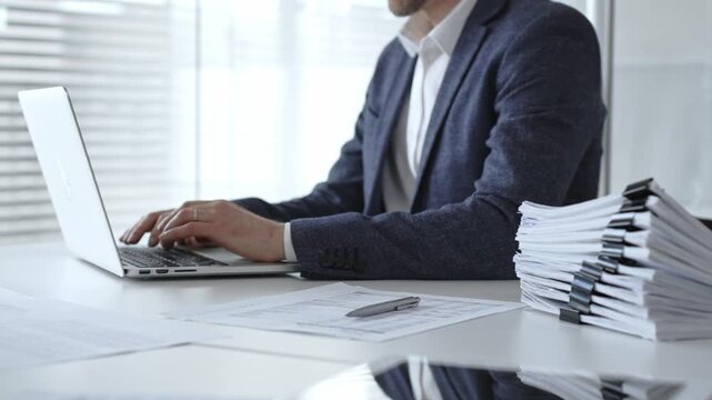 Businessman typing on laptop amid stacked documents and reports, focused on financial data and paperwork in a modern office, conveying productivity, management, and analysis. Audit and taxes
