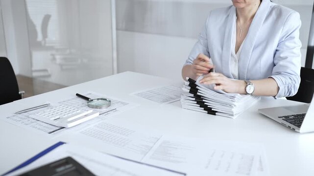 Woman's hands signing and checking financial documents, surrounded by a high stack of paperwork, a calculator, and a laptop, symbolizing busy office work, auditing, and bureaucracy. Audit and taxes