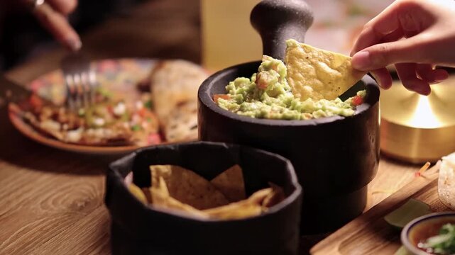 Close up of person dipping tortilla chip into fresh guacamole, enjoying Mexican food at table.