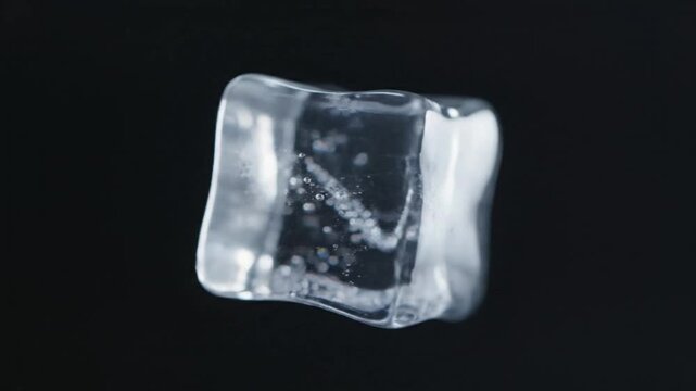 Close-up of a crystal clear ice cube with air bubbles floating and rotating slowly against a solid black background.