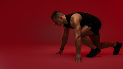 Fototapeta premium Athletic man crouched in explosive sprint starting position against red studio background