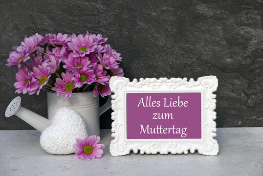 Vase mit pinken Blumen, wei&szlig;e Herz-Deko und wei&szlig;er Bilderrahmen mit Text 'Alles Liebe zum Muttertag' auf grauem Untergrund.