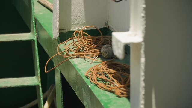 Coiled orange twine on green railing, sunlit closeup of tangled nylon line resting beside white metal bulkhead on ferry deck, practical storage and worn paint textures, industrial maritime vibe