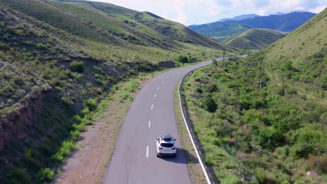 Drone tracking shot follows car driving along winding road near Zhalanash village toward Saty, surrounded by green hills and Tien Shan foothills in southeastern Kazakhstan landscape