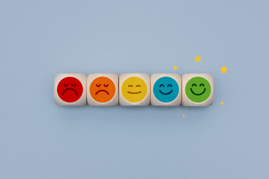 Horizontal row of wooden cubes with colored emoticons from red dissatisfied to green happy with gold stars. Concept net promoter score survey, client feedback experience and reputation management.