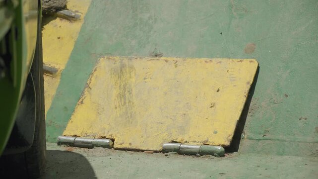 Yellow metal plate on green deck showing worn hinge and textured paint quiet industrial scene with parked vehicle shadow, maintenance vibe, dock worker inspection implied, sunlit gritty surface
