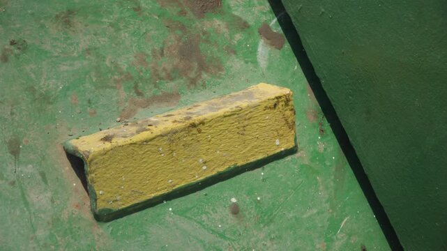Yellow metal block on green surface, closeup showing chipped paint, rust spots, shadowed edge and textured grime, industrial hazard marker for maintenance and inspection, strong contrast and gritty