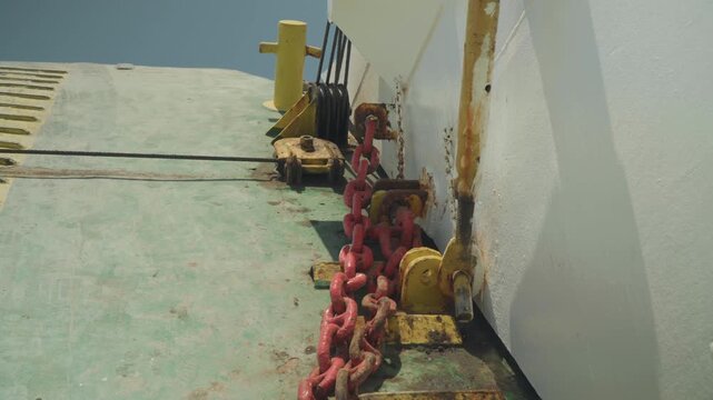 Rusted red chain on green deck, closeup view of bow area with yellow bollard and white hull edge, textured paint and oxidation details, deckhand inspecting mooring hardware, bright daylight and gritty