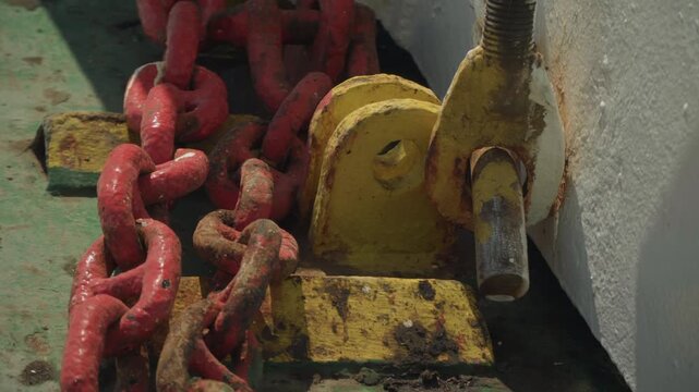 Closeup red chain and yellow pulley on green deck, heavy metal textures with rust spots and flaking paint, mechanical rigging detail showing bracket and shaft, engineer adjusting winch assembly