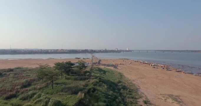 Aerial view of the bustling Kampong Cham riverbank during the Khmer New Year, revealing a bamboo bridge, a crowded sandy beach with local stalls, and the distant city skyline over the Mekong river