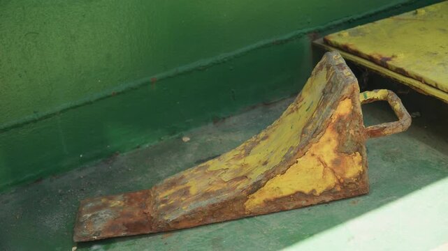 Rusty yellow wheel chock on deck with flaking paint and heavy oxidation, closeup of corroded wedge and loop handle against faded green bulkhead conveys derelict shipyard atmosphere with salvage worker