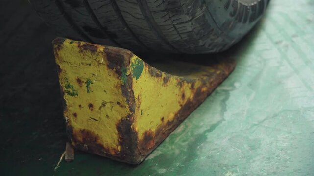 Yellow rusty wheel chock under tire, garage mechanic securing vehicle, corroded metal wedge gripping worn tire, stained concrete floor, textured paint, gritty industrial lighting, maintenance context