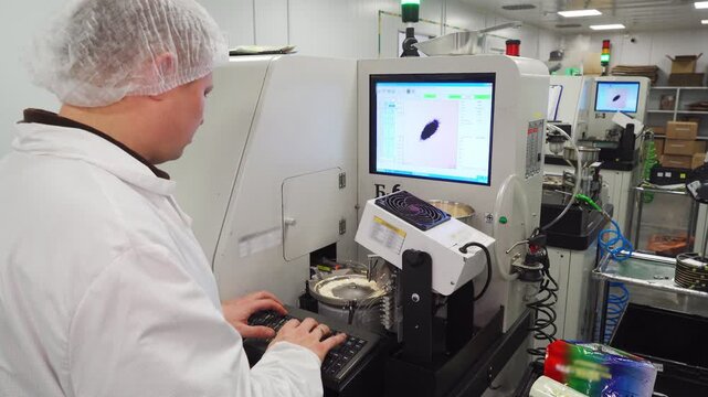 Electronics industry. Industrial software developer programs machine for producing LED lamps. Engineer in a white lab coat works at the keyboard and monitor of a CNC machine in production facility.