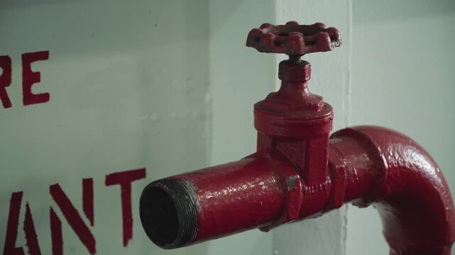 Red fire valve mounted on wall, closeup showing glossy painted pipe and threaded spout, partial emergency signage in background, dim corridor lighting, textured metal with wear and traces