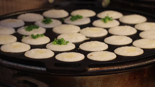 Chef Garnishing Traditional Thai Coconut Pancakes (Khanom Khrok) with Chopped Green Onions on a Cast Iron Griddle