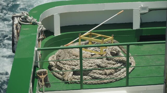 Coiled rope on green ship deck, sunlit weathered rope coil beside yellow bracket and metal railing, ocean spray at stern, mooring readiness vibe, maintenance detail for maritime stock footage