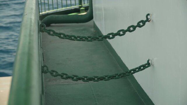 Green ship deck with chains, weathered paint and metal railing, white hull edge and ocean backdrop, mooring hardware like bollard and stanchion visible, industrial texture and calm horizon, close