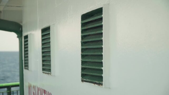 Ship exterior ocean horizon closeup ventilation, green louvered vents set into white painted metal wall, subtle rust at edges, glimpse of green railing and open sea in background, soft cinematic light