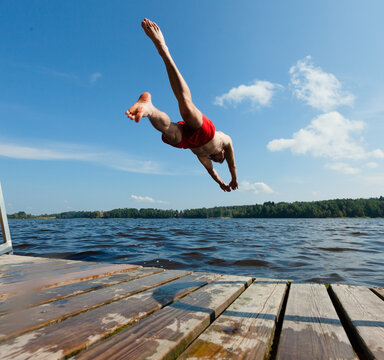 Young man dives off pier into lake captured in mid-air on sunny summer day. Guy in red swim trunks plunges with athletic precision outdoors. Diver descends toward water with powerful form and splash