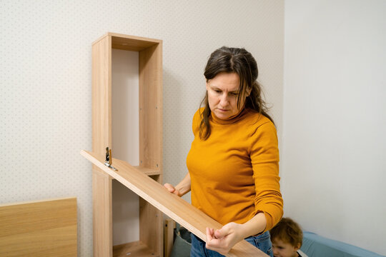 A woman looks thoughtfully at a cabinet door, considering how to correctly attach and mount it. A moment of problem-solving during independent furniture assembly.