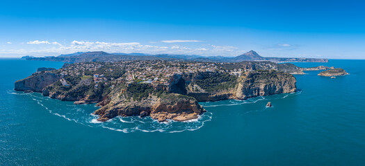 Obraz premium Panoramic view of Costa Blanca coastline in Spain, featuring dramatic rocky cliffs, turquoise Mediterranean Sea, hillside town, and distant mountains under blue sky