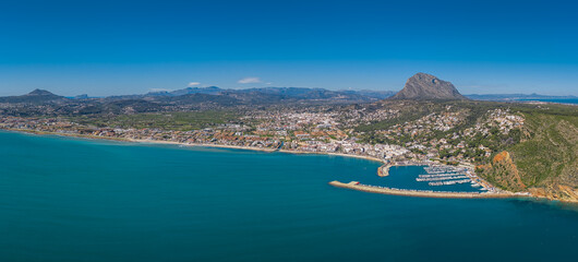 Fototapeta premium Panoramic aerial view of Javea, Costa Blanca, Spain, showing turquoise Mediterranean sea, marina with boats, white town buildings, and iconic Montgo mountain backdrop
