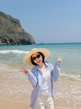Woman enjoys a sunny day at the beach while wearing a straw hat and sunglasses in front of clear blue water and rocky cliffs