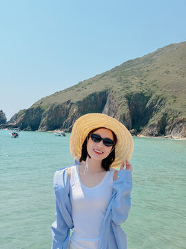 Woman enjoys sunny day at beach wearing sun hat and sunglasses near water with boats and mountains in the background