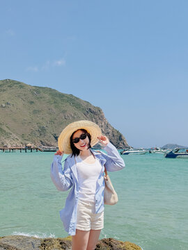 Woman stands on a rocky beach near boats and mountains under blue sky on a sunny day, enjoying the warm weather and scenic view
