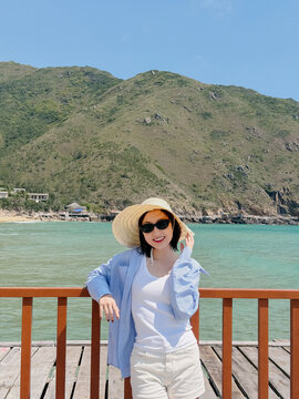 Woman enjoys sunny day on dock by water with mountains in background, wearing sunglasses and straw hat while posing for picture