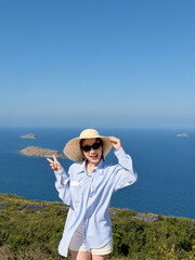 Young Asian woman stands on hilltop near ocean, wearing a sun hat and smiling with peace sign while enjoying sunny day © Huyn