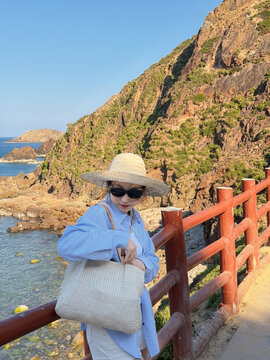 Person in a straw hat stands by a red railing near the coast with rocky cliffs and blue sea in the background during daylight