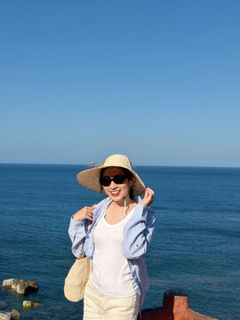 Sunny day at the seaside with an Asian woman smiling and enjoying the view of the blue ocean and clear sky while wearing a sun hat