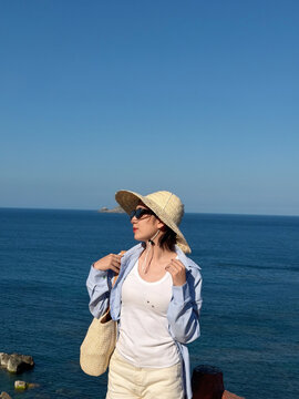 Woman stands by the water wearing sun hat and sunglasses while enjoying the sunny day at the beach near the ocean during afternoon hours