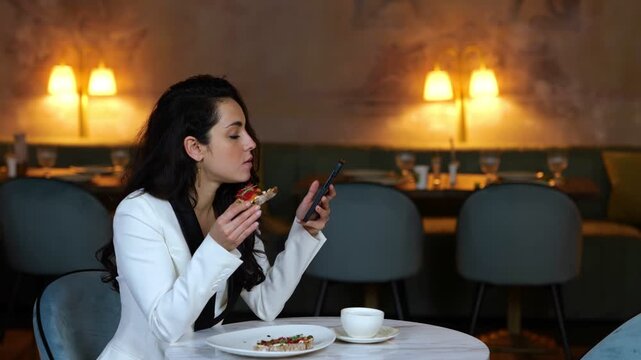 Young lady sitting in restaurant while biting bruschetta and reading a message, holding mobile phone. Copy space. Business, lifestyle concept. Real time video.