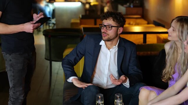 Young man in suit sitting with his friends in the restaurant while ordering from the waiter. Lifestyle and friendship concept. Real time.