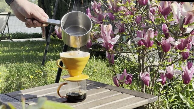Hand pouring hot water from metal pot over pour over coffee dripper on glass cup in sunny garden, blooming magnolia tree in background, slow coffee ritual, outdoor lifestyle and relaxation concept