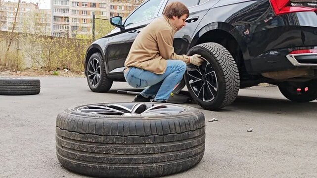 A man changes a tire on a black car outdoors. Seasonal tire change, car maintenance, auto repair.